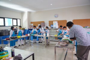 Mengenakan apron biru, siswa tampak sedang bersiap-siap di meja yang disusun menyerupai huruf U