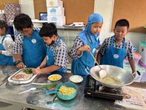 Para siswa saling bekerja sama dan berbagi tugas saat memasak, ada yang menggoreng, ada yang mengiris bahan